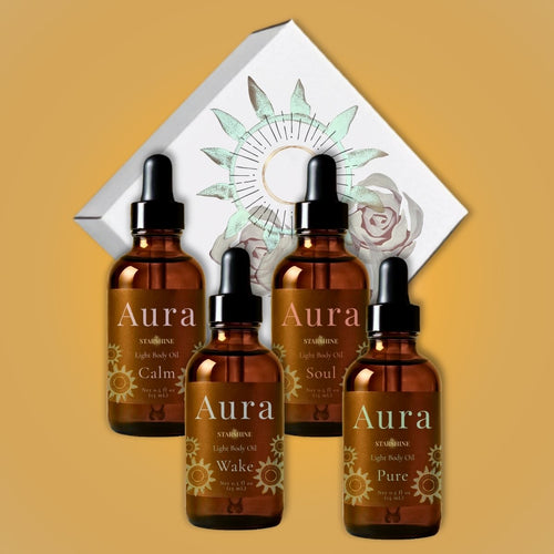 Four amber glass dropper bottles of Aura skincare light body oils arranged in front of a white decorative box with floral and sun illustrations on a warm tan background. scents are frankincense, lavender, peppermint +eucalyptus, and tea tree.