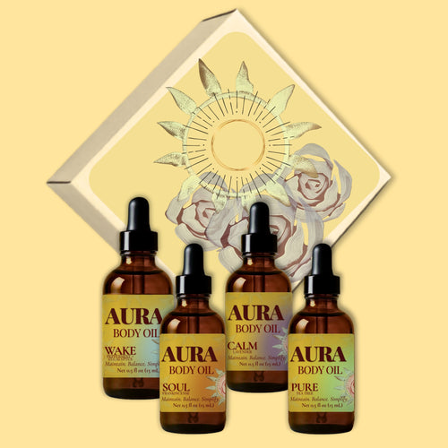 Four amber glass dropper bottles of Aura Body Oil arranged in front of a decorative cream gift box with a sun and rose illustration on a warm beige background. The bottles are labeled Calm Lavender, Wake Eucalyptus + Peppermint, Soul Frankincense, and Pure Tea Tree.