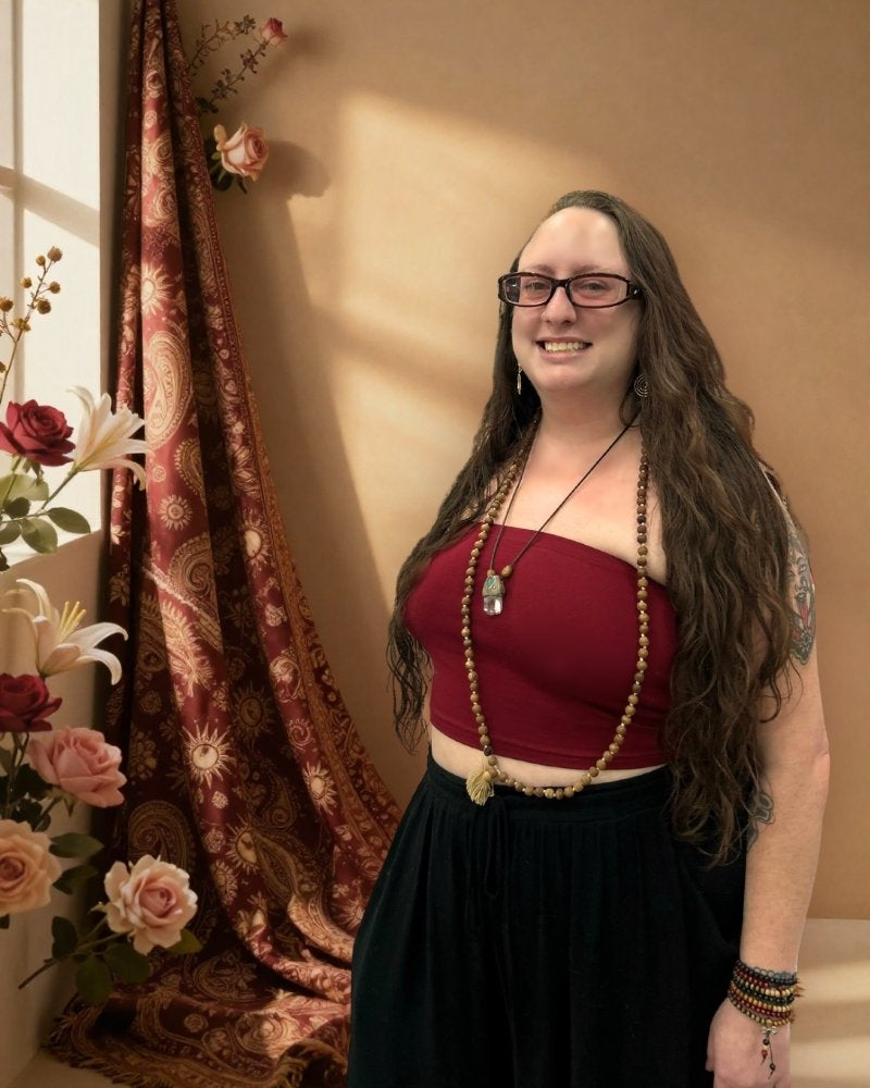 A woman with long wavy brown hair and glasses stands indoors wearing a sleeveless dark red top and a long black skirt. She is accessorized with layered beaded necklaces and bracelets.