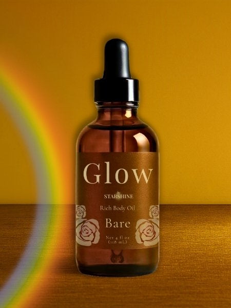 A bottle of Glow bystarsHINE Rich Body Oil bare unscented, featuring an amber glass dropper bottle with a black cap, set against a warm yellow background with a rainbow light leak on the left.