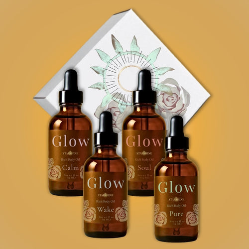 Four amber glass dropper bottles of Glow body oils arranged in front of a white decorative box with a sun and rose illustration on a warm tan background. Scents are frankincense, lavender, peppermint and eucalyptus, and tea tree.