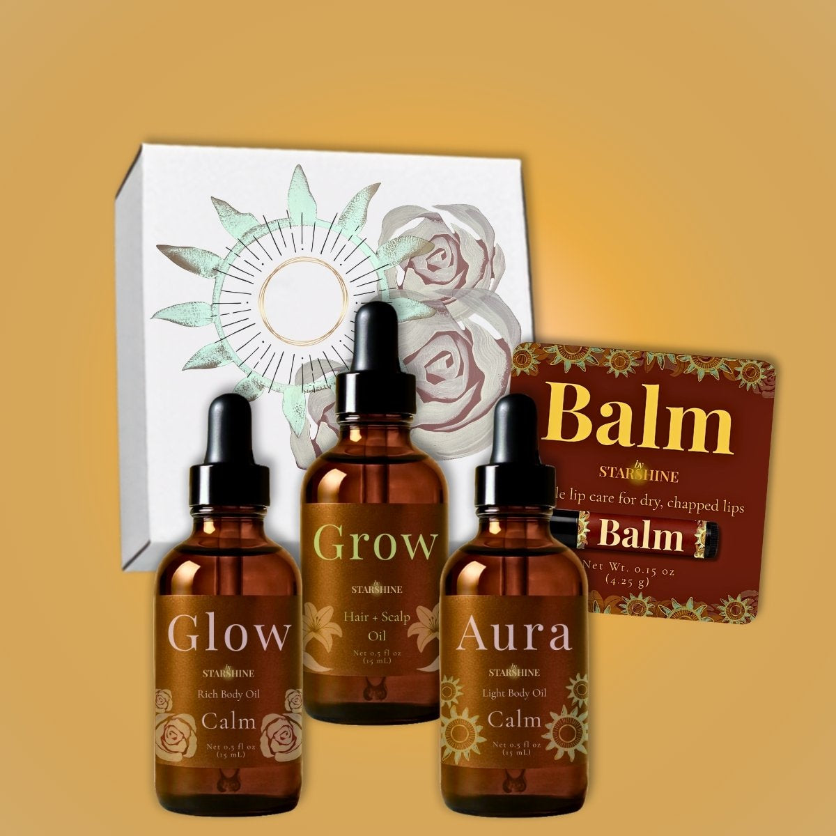 Three amber glass dropper bottles labeled Glow, Grow, and Aura lavender scented body oil for dry skin with black caps are arranged in front of a white gift box with floral artwork, alongside a brown balm packet labeled Balm.