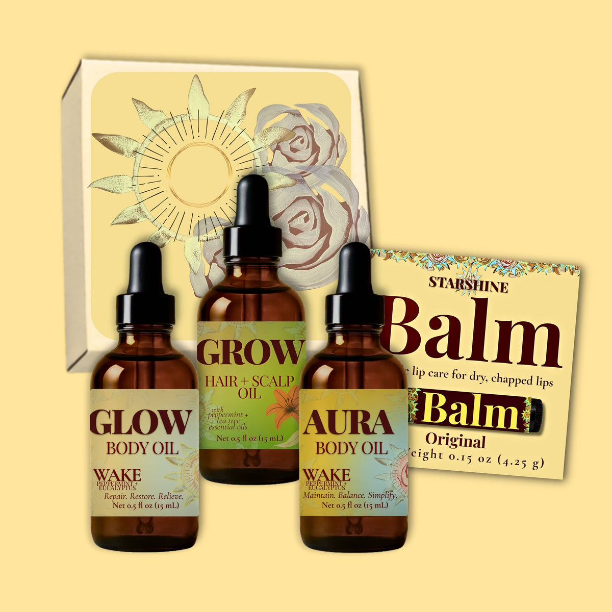 Three amber glass dropper bottles labeled Glow, Grow, and Aura body oils for dry skin peppermint and eucalyptus scented with black caps are arranged in front of a white gift box with floral artwork, alongside a brown balm packet labeled Balm.